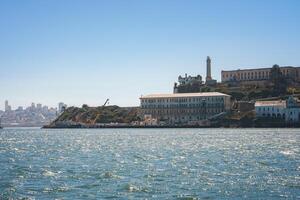 View of Alcatraz Island in San Francisco Bay, California, USA on a clear day. Iconic buildings and lighthouse against a blue sky, with cityscape in the background. photo