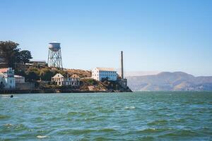 Scenic view of Alcatraz Island in San Francisco Bay, California, USA. Features former prison building, water tower, and coastal hills under blue sky. Peaceful waters. photo