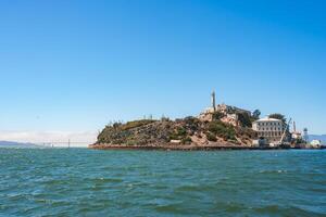 Clear view of Alcatraz Island in San Francisco Bay, California, USA. Features the prison facility, lighthouse, and rugged terrain. Golden Gate Bridge in the background. Sky is clear over calm waters. photo