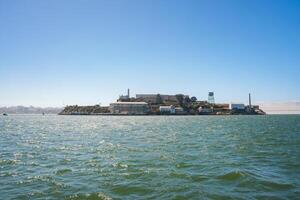 Scenic view of Alcatraz Island in San Francisco Bay on a sunny day. Shows the former prison complex with main cellhouse, water tower, and skyline in the distance. photo