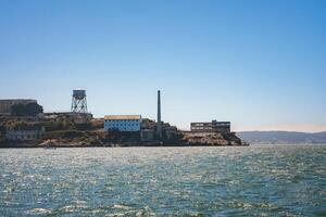 View of Alcatraz Island on a clear day in San Francisco Bay. Features water tower, buildings, smokestack, and boat, capturing former prison's history. photo