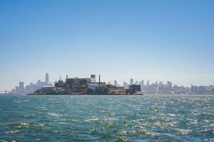 Scenic view of Alcatraz Island in San Francisco Bay under a clear sky, surrounded by old prison structures, guard towers, with city skyline in the background. photo