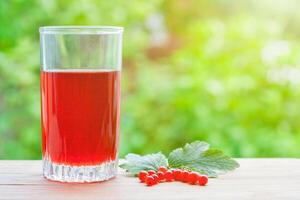 View on a glass with redcurrant juice and berries with leaves on a wooden board illuminated by sunlight on a background of green garden in bokeh. Summer concept photo