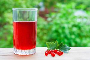 A glass with red juice and branch of redcurrant with leaves on a wooden table on a background of green garden in blur with copy space photo