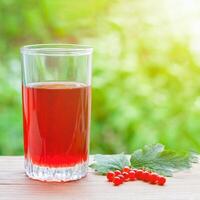A glass with red currant juice and berries with leaves on a wooden table on a background of green bushes in bokeh, illuminated by sunlight photo
