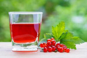 View on a glass with red juice and a branch of redcurrant berries with leaves on a wooden board on a background of green garden in bokeh. Summer concept photo