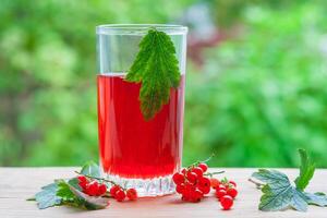 A glass with red currant compote and berries with leaves on a wooden table on a background of green garden in blur photo