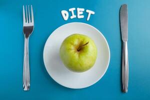 Top view of a green apple on a saucer, with a fork and knife on a blue background with an inscription diet. Concept photo