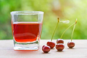 Glass with red cherry juice and a berries on a wooden table, illuminated by sunlight on a background of green garden in bokeh. Summer concept photo
