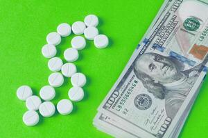 Top view of a white pills are laid out in the shape of a dollar symbol and a heap of banknotes near on a green background photo