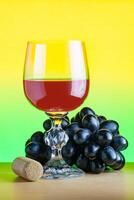A glass of red wine next to bunch of black grapes and a cork on a wooden table on a yellow green gradient background photo