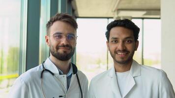 Portrait of two multiracial doctors smiling and looking at camera. Indian and caucasian smiling doctors in white coat working in hospital and looking at camera video