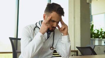 Stressed frustrated doctor wearing white uniform with headache sitting alone at work desk in hospital. Sad young gp feeling desperate and guilty for medical failure video
