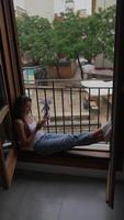 A woman is peacefully relaxing by an open window with a smartphone in an urban setting video
