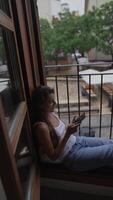 A young woman is relaxing by a window in a casual urban scene, using a smartphone video
