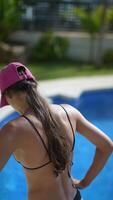 A woman in a cap by a pool, surrounded by greenery, relaxing in the sun at an outdoor setting video