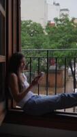 A young woman is peacefully relaxing on a window sill in an urban setting while using her smartphone video