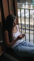 Young woman relaxing and using a smartphone by a window, representing a calm and modern lifestyle video