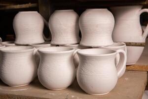 Freshly made clay mugs, pots and bowls drying up on wooden shelves in pottery workshop. Handmade ceramics manufacture. Different clay pots in workshop. photo