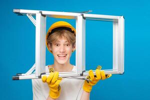 Happy repairman teen in white t-shirt and gloves with yellow helmet in head, holding ladder in hands standing in studio on blue background. A future architect isolated on a blue background photo