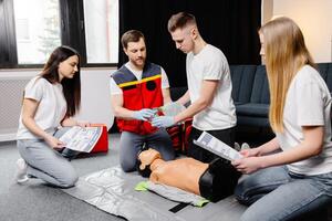 Young man instructor helping to make first aid heart compressions with dummy during the group training indoors photo