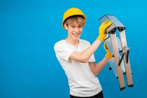 Young repairman teen in white t-shirt and gloves with yellow helmet in head, holding ladder in hands standing in studio on blue background. A future architect isolated on a blue background photo