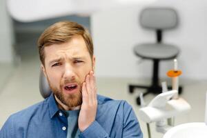 Sad man sitting in dental chair suffering from acute tooth pain, rubbing his cheek, close up photo