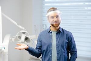 Portrait of handsome male dentist wearing in big protection medical shield on face preparing for dental surgery. Man smiling and looking at camera. photo