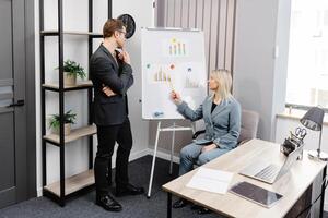 Confident business coach gives presentation on flip chart. A young woman and a man are talking and discussing business project schedules in the office photo