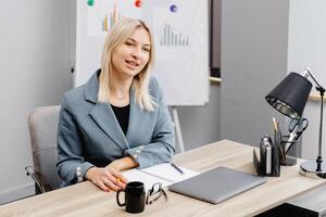 Young business woman sitting at table and taking notes in notebook.On table is laptop and cup of coffee. Student learning online. Blogger. photo