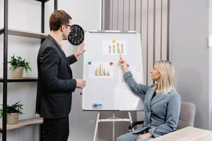 Confident business coach gives presentation on flip chart. A young woman and a man are talking and discussing business project schedules in the office photo