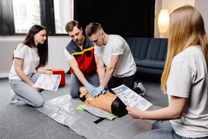 Young man instructor helping to make first aid heart compressions with dummy during the group training indoors photo