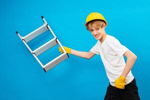 Happy repairman teen in white t-shirt and gloves with yellow helmet in head, holding ladder in hands and running in studio on blue background. A future architect isolated on a blue background photo