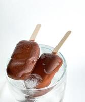 Two popsicle chocolate covered ice cream bars on wooden sticks placed in glass bowl with ice cubes. Top view of frozen treats with condensation on white background. photo