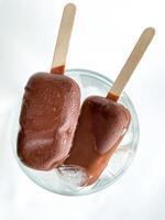 Two popsicle chocolate covered ice cream bars on wooden sticks placed in glass bowl with ice cubes. Top view of frozen treats with condensation on white background. photo