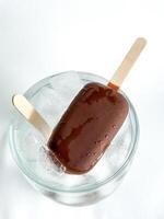 Two popsicle chocolate covered ice cream bars on wooden sticks placed in glass bowl with ice cubes. Top view of frozen treats with condensation on white background. photo
