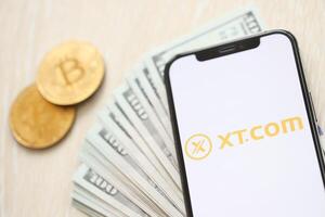 KYIV, UKRAINE - MARCH 15, 2024 XTcom logo on iPhone display screen with dollars and bitcoins on table photo