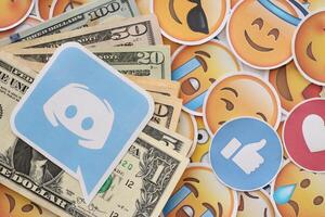 KYIV, UKRAINE - APRIL 20, 2024 Discord logo and many usd money bills. Symbol of famous social network platform and dollar currency banknotes on lot of emoji reactions photo