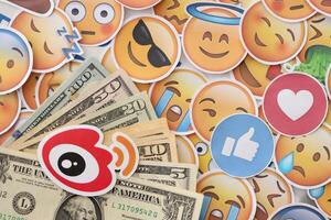 KYIV, UKRAINE - APRIL 20, 2024 Weibo logo and many usd money bills. Symbol of famous social network platform and dollar currency banknotes on lot of emoji reactions photo