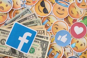 KYIV, UKRAINE - APRIL 20, 2024 Facebook logo and many usd money bills. Symbol of famous social network platform and dollar currency banknotes on lot of emoji reactions photo