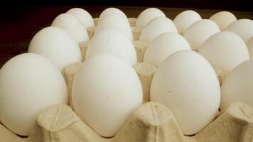 A close up shot of white chicken eggs in a cardboard carton. The eggs are arranged in a row and the carton is made of brown cardboard. The is shot in a dark studio with a black background. video