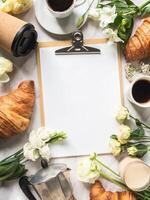 Flat lay frame of cups of coffee, flowers, croissants and clipboard with blank sheet for menu photo