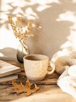 Cozy autumn home still life of dry flowers in vase, mug with drink, a knitted blanket and magazines on a rustic wooden table and shadows on the wall from the bright setting sun photo