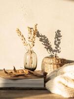 Cozy autumn home still life of dry flowers in vase, straw box, knitted blanket and magazines on a rustic wooden table and shadows on the wall from the bright setting sun photo