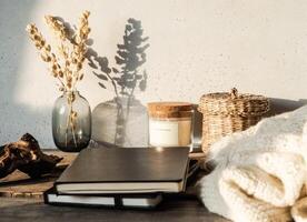 Cozy autumn home still life of dry flowers in vase, straw box, glass candle, knitted blanket and black notepads on a rustic wooden table and shadows on the wall from the bright setting sun photo