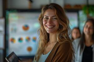 A young woman with long blonde hair smiles confidently as she holds a tablet computer in an office setting. She is standing in front of a whiteboard with charts and graphs visible in the background photo