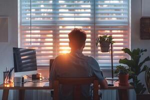 Man working at desk with laptop facing sunset through window blinds photo