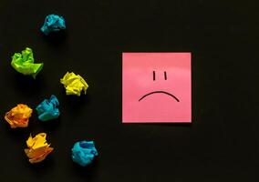upset emoji and crumpled sheets of colored paper on a dark background, failed attempts, motivation photo