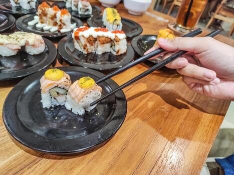 Hand holding chopsticks preparing to take sushi on a plate on a table in a Japanese restaurant with lots of sushi in the background photo