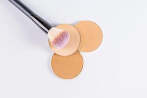 Three round blocks of compact powder in different tones, a large makeup brush lies on them. top view. white background. a layout product photo